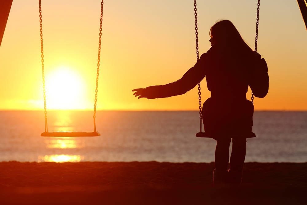 Woman sitting on a swing reaching out for empty swing with sunset in background